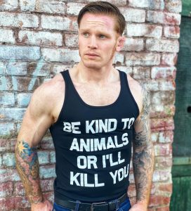 Man with tattoos against wall wearing black be kind to animals or i'll kill you tank top