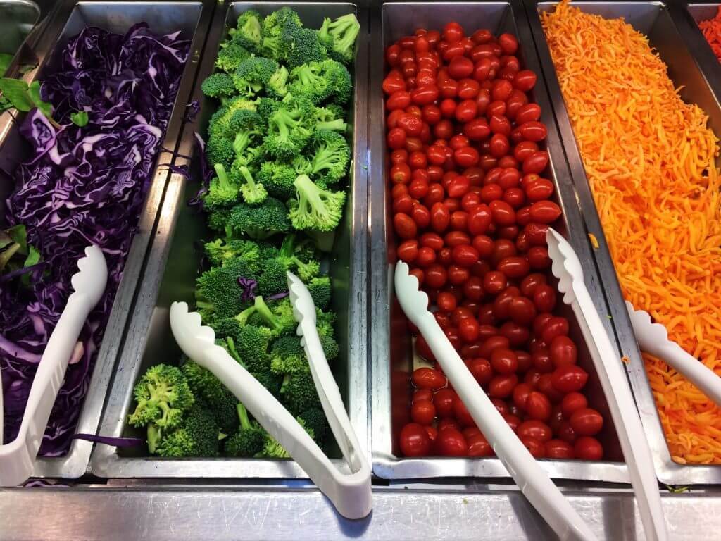 salad bar with sliced red cabbage broccoli tomatoes and cheese in metal inserts with white plastic tongs