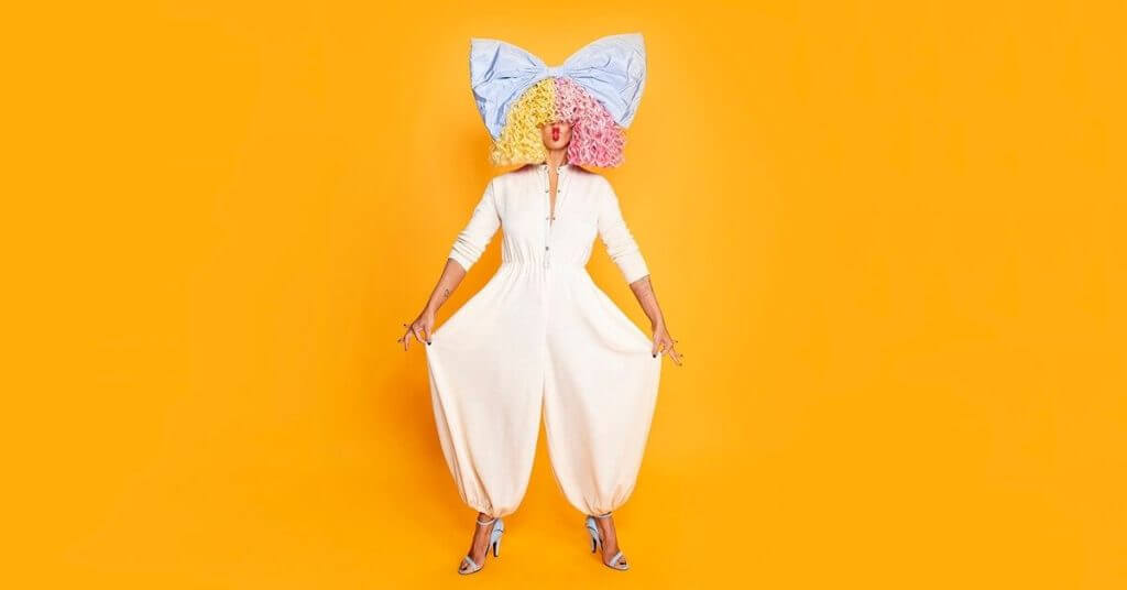 Sia wearing cream humpsuit and large bow on yellow and pink hair against orange background