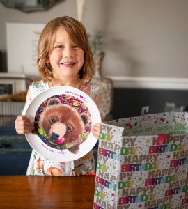 girl holding a be-ve kids plate grandma loves you berry bear next to a happy birthday gift bag